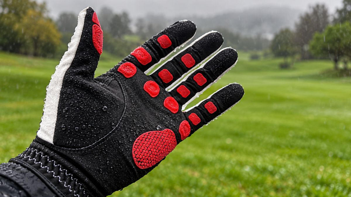 EXO golf glove covered in raindrops, palm facing the camera, showing red Zyntech™ nanocell grip pads on a rainy, misty golf course with a green fairway